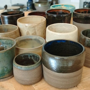 Peut inclure: Une collection de tasses et de petits bols en céramique faits à la main, de différentes tailles et couleurs. La poterie présente une gamme de tons terreux, notamment marron, beige et sarcelle, avec des finitions brillantes et mates.
