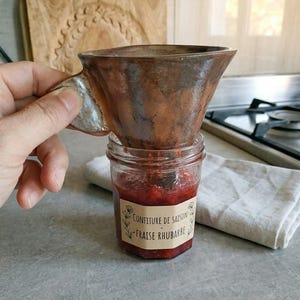 Könnte beinhalten: Ein handgefertigter Keramiktrichter in Braun und Silber wird über einem Glas roter Marmelade gehalten. Das Glas trägt das Etikett "Confiture de saison - Fraise Rhubarbe". Im Hintergrund ist ein Holzschneidebrett zu sehen.
