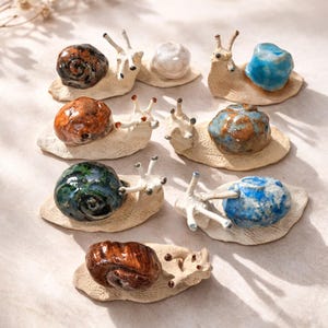 Peut inclure: Une collection de sept petites figurines d'escargots en céramique, chacune dotée d'un corps texturé blanc cassé et d'une coquille brillante aux couleurs distinctes. Les coquilles présentent diverses finitions, dont brun, bleu, vert et blanc.