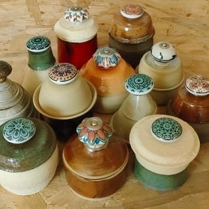 May include: A collection of decorative ceramic jars with lids. The jars vary in size, shape, and color, including beige, brown, green, and red. Each jar is topped with a unique, colorful knob with floral or geometric patterns.