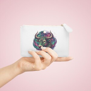 Pode incluir: Bolsa branca com zíper com uma ilustração colorida da cabeça de um touro com grandes chifres, em um fundo cósmico. O touro é verde com detalhes florais. A bolsa tem um puxador branco e é segurada em uma mão.