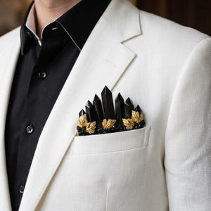 May include: A white blazer with a black shirt. The breast pocket is adorned with a decorative arrangement of black crystal-like shapes and gold-coloured leaf accents, creating a unique and stylish accessory.