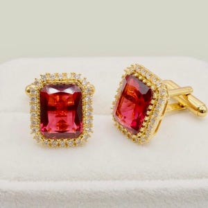May include: A pair of gold-toned cufflinks with large, rectangular, red gemstones. Each gemstone is surrounded by a halo of small, clear crystals. The cufflinks are displayed on a white surface.