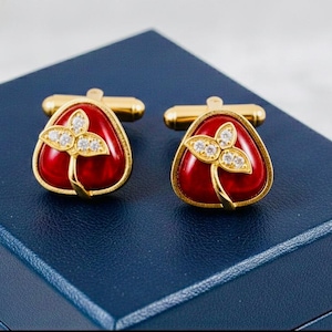 Puede incluir: Un par de gemelos dorados con un centro rojo en forma de lágrima. Cada gemelo presenta un diseño floral dorado con tres cristales transparentes. Los gemelos se muestran en una caja azul oscuro.