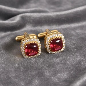 May include: A pair of gold cufflinks featuring square, ruby-red centre stones. Each cufflink is framed by a halo of small, clear gemstones. The cufflinks are presented on a grey, velvety surface.