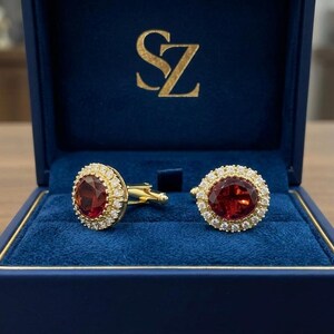 May include: Gold cufflinks featuring large, oval, red gemstones encircled by smaller, clear stones. Presented in a blue velvet box, the cufflinks are inscribed with the gold initials "SZ".