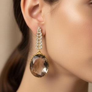 May include: Gold-toned drop earrings featuring a large, faceted, smoky quartz stone. The earrings have a decorative, leaf-like design with clear crystals. The earrings are a statement piece.