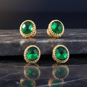Könnte beinhalten: Vier goldfarbene Ohrstecker mit großen, facettierten smaragdgrünen Edelsteinen. Jeder Ohrring hat einen detaillierten, strukturierten Goldrand. Die Ohrringe sind auf einer dunklen, marmorierten Oberfläche angeordnet.