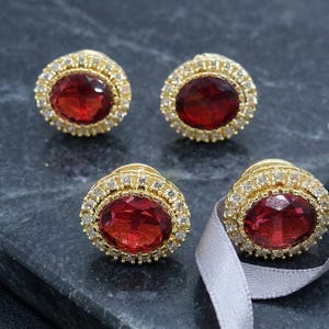 May include: Four gold-toned stud earrings with oval, deep red gemstones. Each earring is surrounded by a halo of small, clear gemstones. A silver ribbon is draped near the earrings, set against a dark, textured surface.