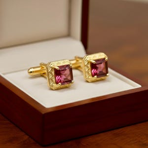 May include: A pair of gold cufflinks, each featuring a square, pink gemstone. The cufflinks are presented in a wooden box with a cream-coloured interior. The gemstones are set in a decorative, textured gold frame.