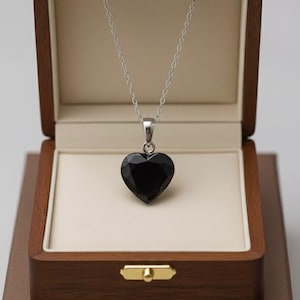 May include: A silver necklace with a black heart-shaped pendant presented in an open wooden jewellery box. The box is lined with cream fabric and has a gold clasp. The box sits on a brown surface.
