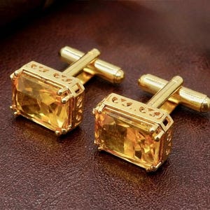 Puede incluir: Un par de gemelos dorados con piedras amarillas rectangulares facetadas engastadas en monturas de metal calado y ornamentado. Cada gemelo cuenta con un mecanismo de cierre de barra en T articulado en la parte posterior para asegurar los puños.