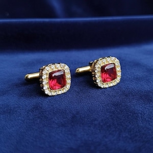 May include: Two gold cufflinks, each featuring a square, deep red gemstone at the centre. The gemstones are framed by a border of small, clear crystals. The background is a dark blue fabric.