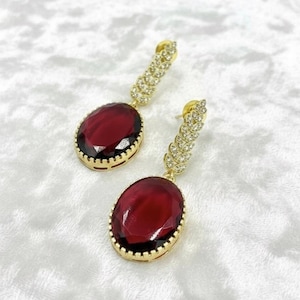 May include: A pair of gold earrings with large, oval, deep red gemstones. The gemstones are set in a gold frame. The earring posts and connecting elements are decorated with small, sparkling crystals, adding glamour to the jewellery.