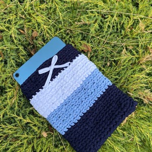 May include: A navy blue, white, and light blue crocheted tablet sleeve. The sleeve has a striped design and a white ribbon closure. The tablet is partially visible, and the sleeve is resting on a bed of green foliage.