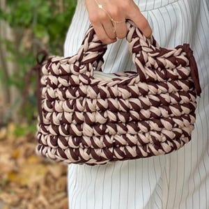 Puede incluir: Un bolso de mano tejido, con un patr&oacute;n repetitivo marr&oacute;n y beige. El bolso tiene forma rectangular con un asa trenzada. El bolso est&aacute; hecho de un material suave y flexible. El bolso lo sostiene una persona.