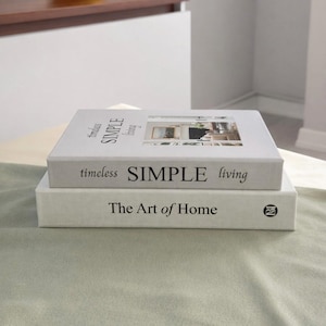 Puede incluir: Dos libros decorativos apilados uno encima del otro. El libro superior es blanco con las palabras "timeless SIMPLE living" impresas en la portada. El libro inferior también es blanco y dice "The Art of Home".