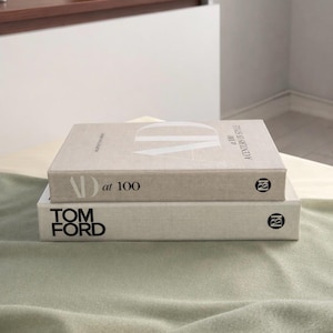 Puede incluir: Dos libros de tapa dura apilados uno encima del otro. El libro superior se titula "AD at 100 A Century of Style" con un gran gráfico "AD". El libro inferior se titula "TOM FORD". Ambos libros tienen una combinación de colores neutros.