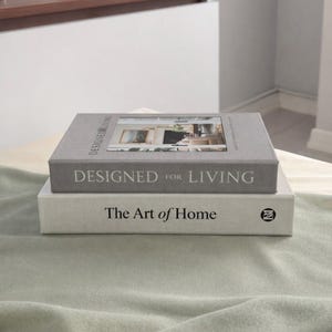 Può includere: Due libri rilegati impilati. Il libro superiore è grigio e recita "DESIGNED FOR LIVING" con un'immagine di una stanza. Il libro inferiore è bianco e recita "The Art of Home".