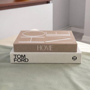 Può includere: Due libri decorativi impilati uno sopra l'altro. Il libro superiore è beige con la parola "HOME" in bianco. Il libro inferiore è bianco con le parole "TOM FORD" in nero.