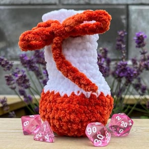 May include: A white and orange crochet potion bottle dice pouch with a bow. The pouch is decorated with a white body and orange accents. The image also shows several pink dice.