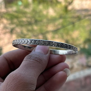 Puede incluir: Una pulsera de plata con un patrón grabado de símbolos repetidos. La pulsera se sostiene en una mano, con un fondo borroso que sugiere un entorno al aire libre. El diseño es intrincado y detallado, mostrando una estética tradicional.