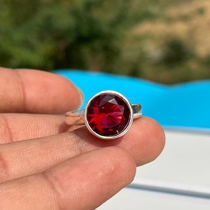 Può includere: Un anello d'argento con una grande gemma rotonda di colore rosso intenso. La gemma è sfaccettata e incastonata in una lunetta. L'anello è tenuto in mano su uno sfondo sfocato.