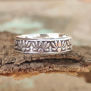 Può includere: Un anello d'argento con un motivo floreale a forma di fiore di loto ripetuto. L'anello è posto su uno sfondo di legno naturale, che mette in risalto l'intricato motivo floreale. La fascia dell'anello è larga circa 0,6 cm.