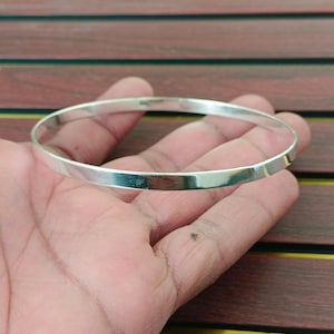 Puede incluir: Un brazalete de plata con una superficie plana y pulida. El brazalete es redondo y parece estar hecho de metal. El brazalete se sostiene en una mano, con una superficie de madera en el fondo.