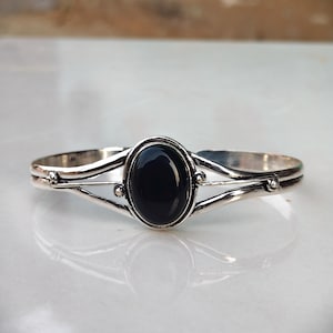 Puede incluir: Un brazalete de plata con una piedra negra ovalada en el centro. La banda metálica se divide en dos brazos delgados a cada lado de la piedra, decorados con pequeñas esferas de plata en los puntos donde los brazos se unen a la montura.