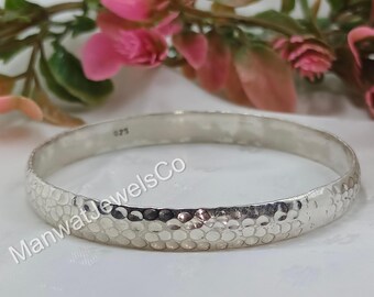May include: A silver bangle bracelet with a hammered texture. The bracelet is round and has a series of small, evenly spaced indentations across its surface. The background includes pink flowers and the text "ManwatJewelsCo".