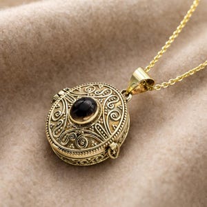 May include: A gold-colored pendant necklace with an oval black stone set in the center. The round pendant features intricate scrollwork and opens to reveal a small compartment. The necklace chain is also gold.