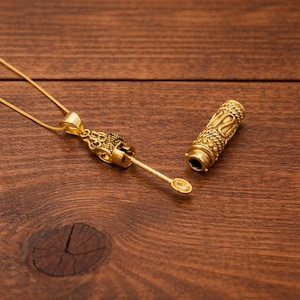 Könnte beinhalten: Eine goldfarbene Halskette mit einem Anhänger mit aufwendigem Design und einem kleinen löffelartigen Aufsatz. Daneben befindet sich ein passender zylindrischer Behälter mit dekorativen Mustern. Die Artikel werden auf einer Holzoberfläche präsentiert.