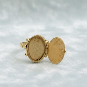 May include: A gold-colored locket ring with an oval-shaped compartment that opens. The ring has decorative scrollwork on the sides of the compartment. The locket is open, revealing the interior.