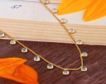 May include: A delicate gold-colored necklace featuring small, round, light-colored gemstones. The necklace has a chain design with evenly spaced gemstones. The background includes a wooden surface and orange flower petals.