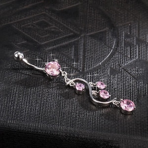 May include: A silver belly button ring with a curved barbell design. The ring features a large pink gemstone at the top and a dangling floral design with smaller pink gemstones. The text "Twinkle Tummy" is in a red box in the upper left corner.