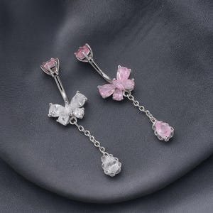 May include: Two silver-toned belly button rings. One features a clear, butterfly-shaped gem and a dangling clear teardrop gem. The other has a pink flower gem and a dangling pink teardrop gem. Both have a curved bar.