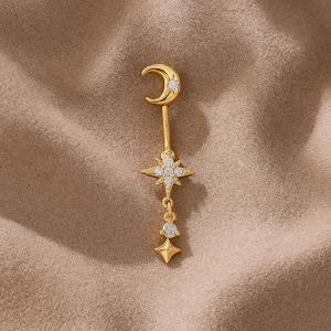 Könnte beinhalten: Goldfarbener Ohrring im Himmels-Thema mit einem Halbmond, einem Stern, einem Tropfen und einem kleinen Stern. Der Ohrring ist mit kleinen, klaren Edelsteinen besetzt. Der Ohrring wird vor einem neutralen, strukturierten Hintergrund präsentiert.