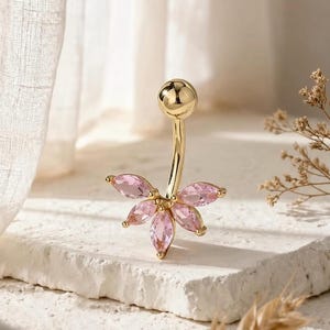 Könnte beinhalten: Ein goldfarbener Bauchnabelpiercing mit floralem Design. Der Ring hat einen gebogenen Stab mit einem kleinen, runden Ball an der Spitze und einer Ansammlung rosa, marquiseförmiger Edelsteine, die eine Blume bilden. Die Edelsteine sind in goldfarbenen Zinken gefasst.
