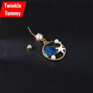 Puede incluir: Un piercing para el ombligo de color dorado con un diseño de concha azul y estrella blanca, acentuado con perlas y una pequeña gema. El texto "Twinkle Tummy" está en un cuadro rojo en la esquina superior izquierda.