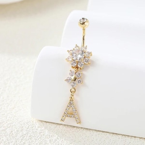 Pode incluir: Anel de umbigo dourado com um design floral e a letra "A" adornada com cristais transparentes. O anel apresenta uma barra curva e uma bola superior brilhante. O texto "Twinkle Tummy" está em uma caixa vermelha no canto superior esquerdo.