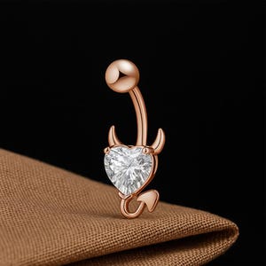May include: Rose gold belly button ring with a heart-shaped, clear gemstone. The ring features a devil-themed design with horns and a tail. The curved barbell has a small ball at the top.
