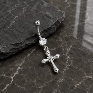 Könnte beinhalten: Ein silberfarbener Bauchnabelpiercing mit gebogener Stange. Er hat einen klaren, runden Edelstein und einen Anhänger in Form eines Kreuzes. Das Kreuz hat ein verziertes Design.