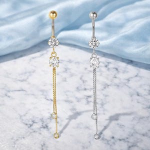 Puede incluir: Dos piercings para el ombligo, uno dorado y otro plateado, cada uno con una cadena colgante con tres gemas redondas y transparentes. La parte superior de cada anillo tiene una pequeña bola redonda. El fondo es de tela azul claro.