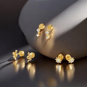 May include: A collection of gold-colored stud earrings. The earrings feature various designs, including dangling crystals, snake shapes, and starbursts. The earrings are displayed on a reflective surface.