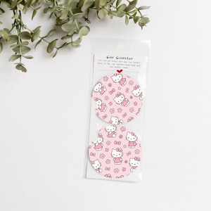 May include: Two pink car coasters with a repeating pattern of a cartoon cat and flowers. The coasters are packaged in a clear plastic sleeve. The text on the packaging reads "Car Coaster".