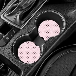 May include: Two pink car coasters with a black polka dot pattern are placed in a car's cup holders. The coasters are round and made of absorbent material. The car's interior is black.