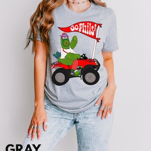 May include: Gray t-shirt featuring a cartoon character riding a red ATV and holding a flag that says "GO PHILS!". The character is green and wearing a baseball cap. The word "GRAY" is printed in large, black letters at the bottom.