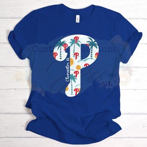 May include: A blue t-shirt featuring a white "P" logo with a tropical design of palm trees, a sun, and the text "Clearwater, FL". The shirt is made of a soft material.