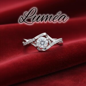 May include: A silver ring with a large, round, clear gemstone at the centre. The band has a swirling design with smaller, clear gemstones. The ring is set against a red background, with the word "Lumea" in cursive script above.